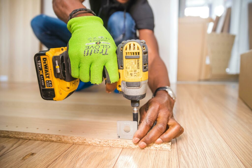 Des mains protégées par des gants verts utilisent une perceuse jaune pour fixer un morceau de bois, représentant un travail manuel précis dans le contexte d'un projet de bricolage ou de rénovation domiciliaire.