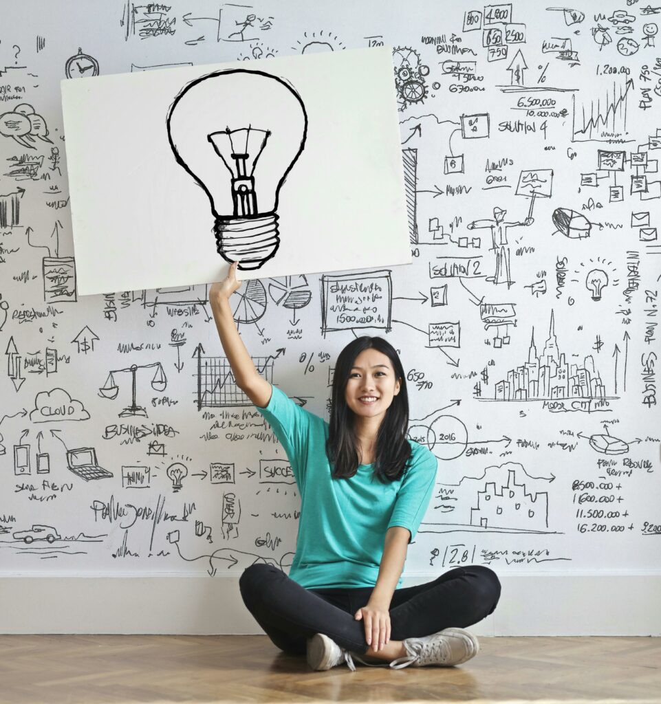 Une femme assise sourit joyeusement en tenant une affiche avec un dessin d'ampoule, devant un mur couvert de croquis et de formules évoquant la créativité et l'inspiration.