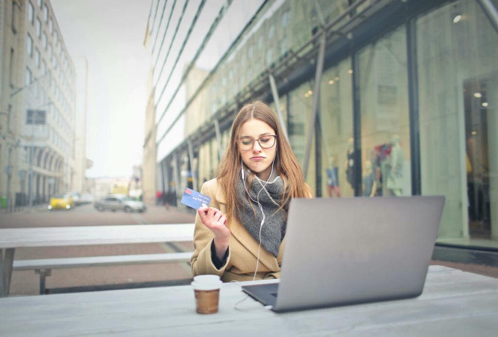 Une jeune femme assise dehors, concentrée sur son ordinateur portable, tient une carte de crédit, illustrant peut-être le shopping en ligne ou la gestion des finances personnelles dans un cadre urbain.
