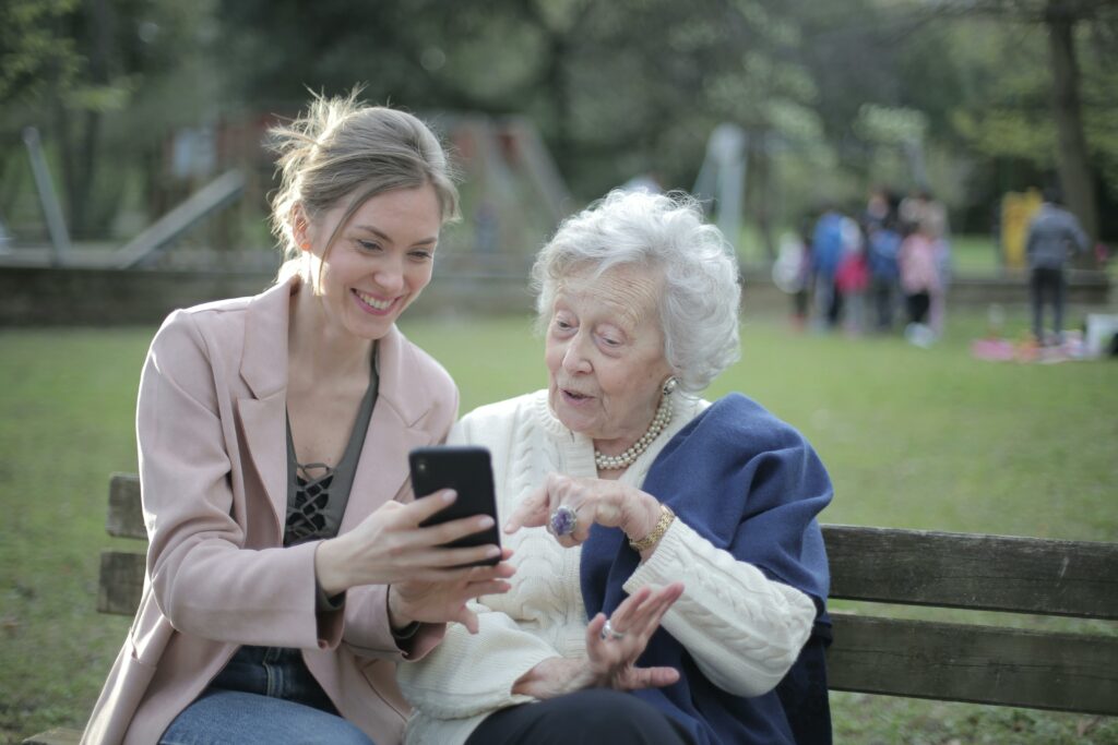 Une femme jeune montre à une personne âgée quelque chose sur un smartphone dans un parc, un moment de partage intergénérationnel.