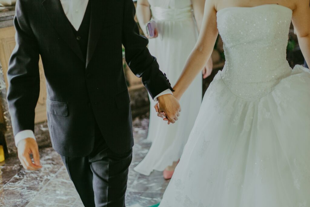 Un couple en tenue de mariage tient les mains, le focus sur leurs mains et les détails des vêtements suggérant un moment romantique et formel.