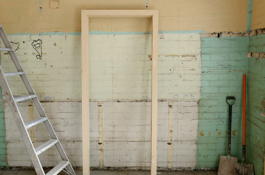 Un cadre de porte en bois érigé dans une pièce en rénovation, avec des murs partiellement dénudés et une échelle à côté, suggérant des travaux d'amélioration ou de réparation de l'habitat.