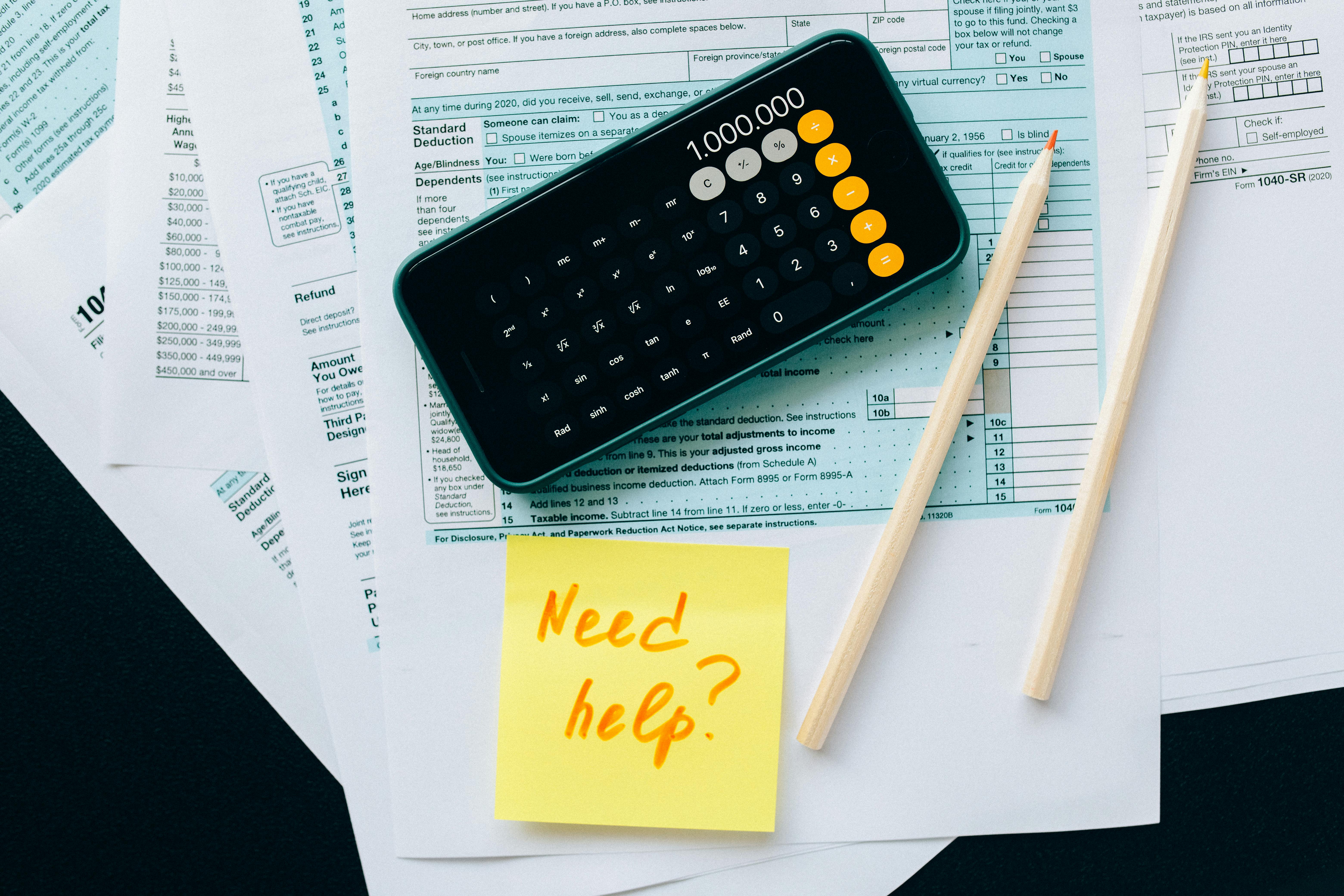 Un calculateur et un stylo posés sur des documents fiscaux avec un post-it disant "Need help?", ce qui suggère la préparation de déclarations fiscales ou la gestion financière.