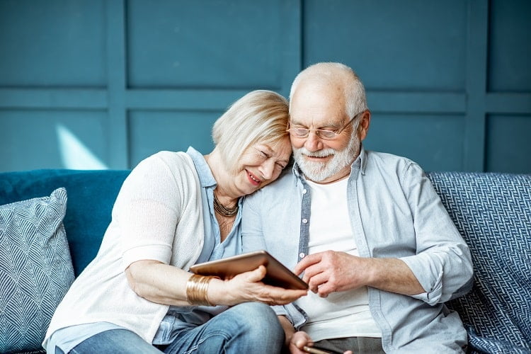 Un couple âgé partage un moment de complicité en regardant une tablette numérique ensemble, confortablement assis sur un canapé.