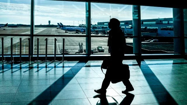Silhouette d'une personne marchant dans un aéroport avec des avions visibles à travers les fenêtres, évoquant le voyage ou le transit.