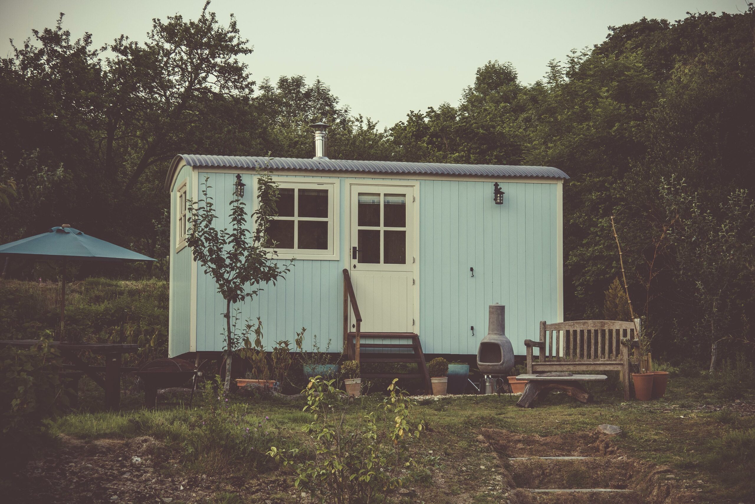 Une petite maison bleue à la campagne avec un jardin négligé et divers objets ménagers à l'extérieur, évoquant un cadre rustique et isolé.