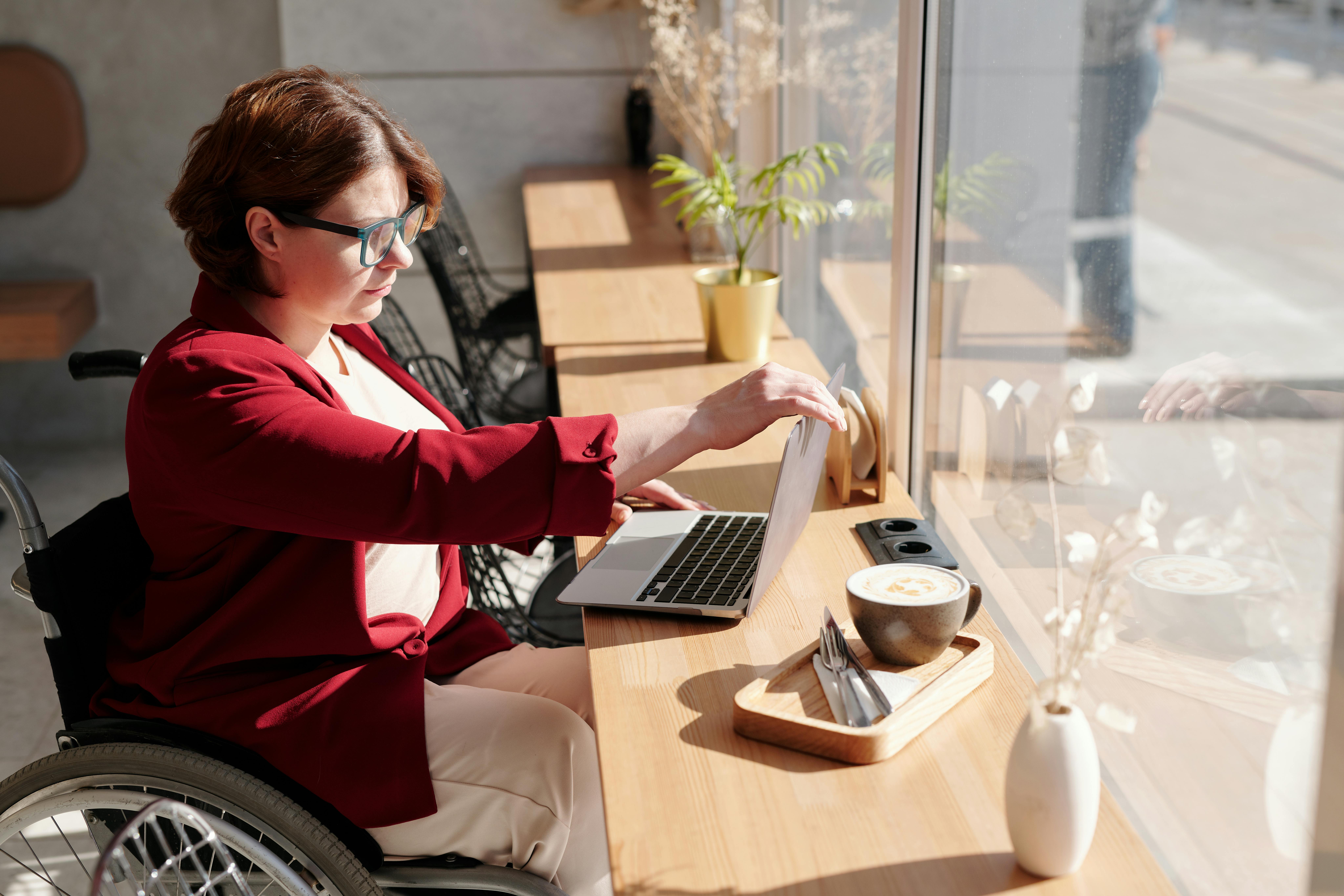 Une femme en fauteuil roulant travaillant sur un ordinateur portable dans un café, incarnant l'indépendance et la normalité de la vie professionnelle pour les personnes handicapées.