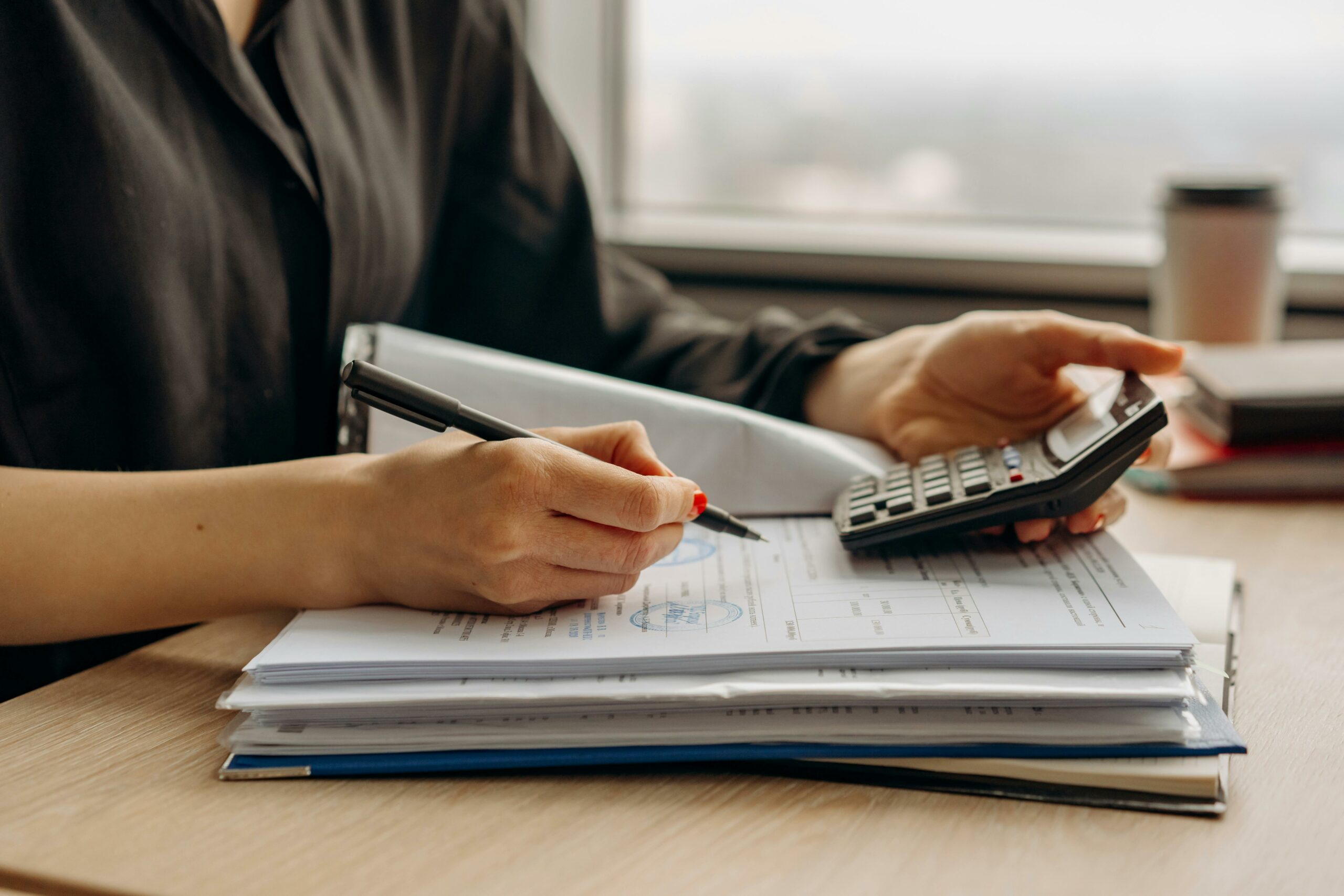 Une femme utilise une calculatrice et écrit des notes sur des documents, dans un bureau lumineux, indiquant un travail de comptabilité ou de budget.