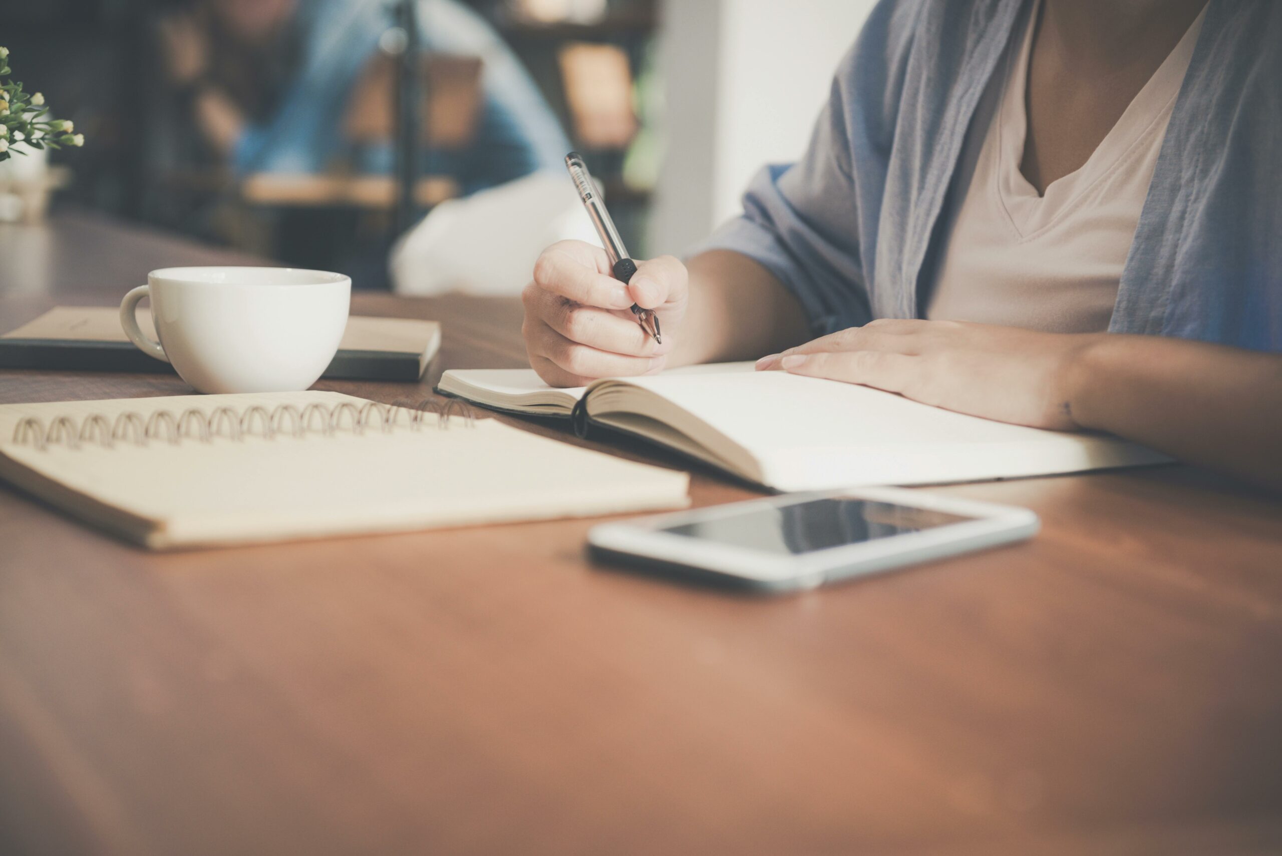 Une personne est en train d'écrire dans un cahier sur une table en bois, à côté d'une tasse de café et d'un téléphone portable, une scène qui évoque la concentration et la réflexion.