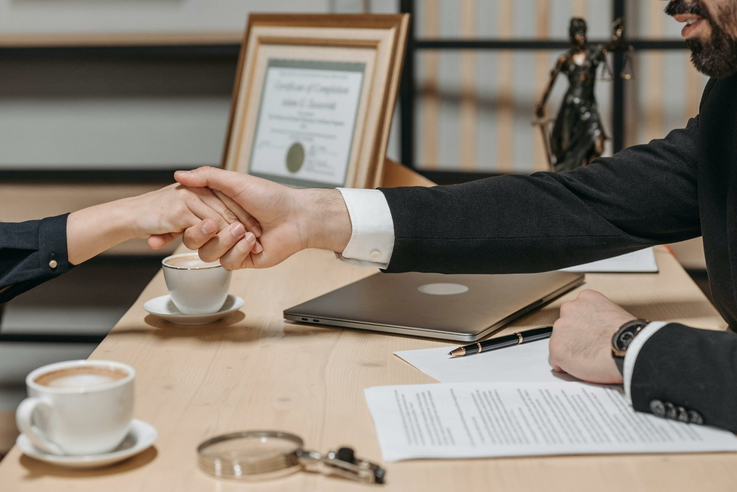 Une poignée de main entre deux personnes assises à une table, avec des documents et une tasse de café devant eux.