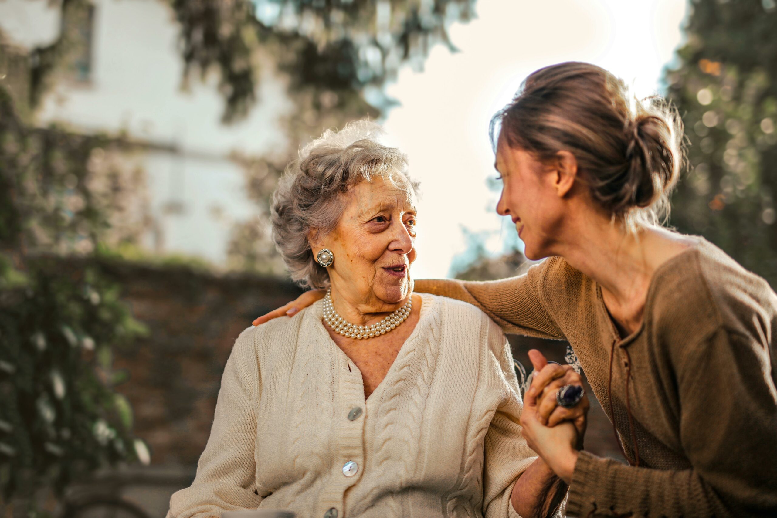 Une femme âgée souriante, portant un collier de perles, est assise et regardée affectueusement par une femme plus jeune qui lui tient la main et lui touche l'épaule, dans un jardin ensoleillé.